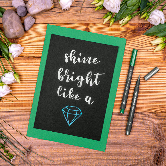 Motivational card with 'shine bright like a' text on a wooden surface with flowers and pens.