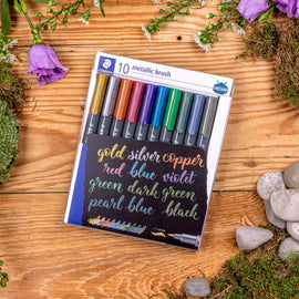 Set of metallic brush pens in a package on a wooden surface with flowers and stones.