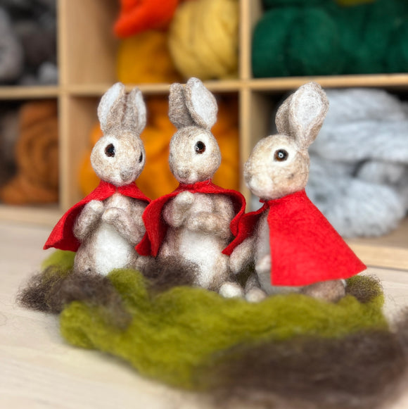Three felted rabbits wearing red cloaks on a green fabric with yarn balls in the background.