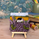 Decorative candle with a landscape design featuring a house, lavender, and a mountain on a wooden stand.