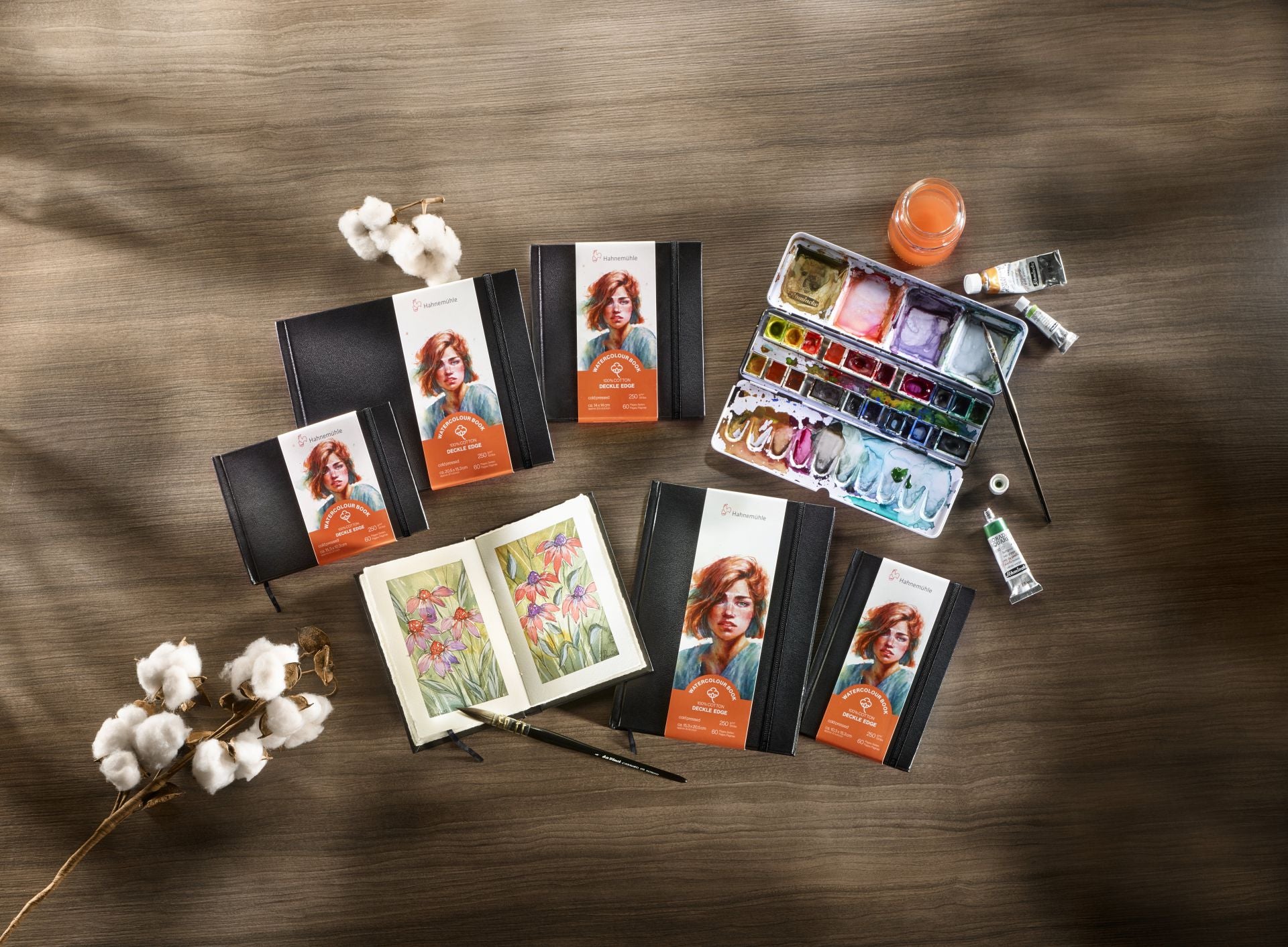 Art supplies including watercolor sets and books on a wooden surface with cotton branches.